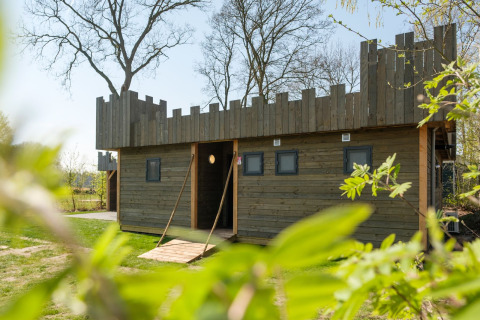 Holzhütte mit Burgmotiv im Kampeerdorp de Zandstuve in den Niederlanden, umgeben von Grün und Bäumen.