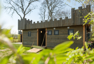 Casa de madera con temática de castillo en Kampeerdorp de Zandstuve, Países Bajos, rodeada de vegetación y árboles.