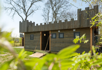 Holzhütte mit Burgmotiv im Kampeerdorp de Zandstuve in den Niederlanden, umgeben von Grün und Bäumen.