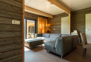 Moderne lodge med trævægge, gråt hjørnesofa og store vinduer på Castle Uylenburght, Kampeerdorp de Zandstuve.