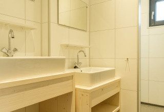 Baño moderno con dos lavabos blancos, muebles de madera clara y espejos en una pared de azulejos.