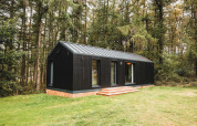 Zwarte houten Koya Tiny Cabin lodge in het bos bij Wilsumer Berge, Duitsland, met grasveld eromheen.