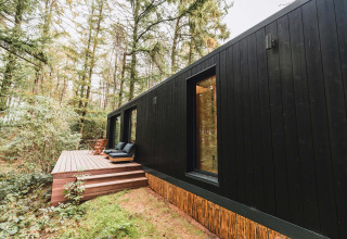 Koya Tiny Cabin negra en Wilsumer Berge, Alemania, rodeada de bosque y con terraza de madera y asientos.