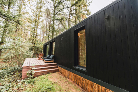 Koya Tiny Cabin negra en Wilsumer Berge, Alemania, rodeada de bosque y con terraza de madera y asientos.