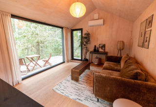 Sala moderna en Koya Tiny Cabin con ventanales grandes, sofá marrón y vista al bosque en Wilsumer Berge, Alemania.