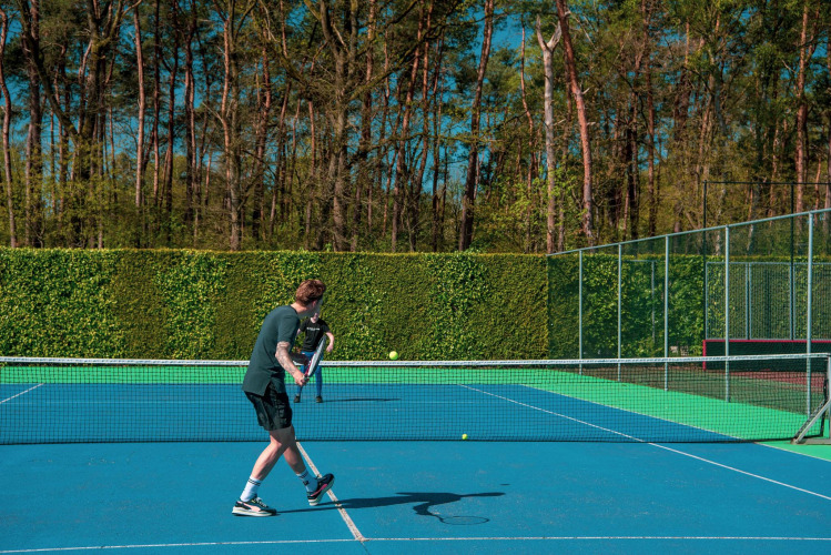 Man playing tennis on a sunny day at Pavilion 6, De Achterhoek lodge, Netherlands, surrounded by greenery.