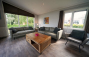 Woonkamer in L-Pavilion 6 te De Biesbosch, Nederland, met grote ramen, zitbanken en houten tafel.