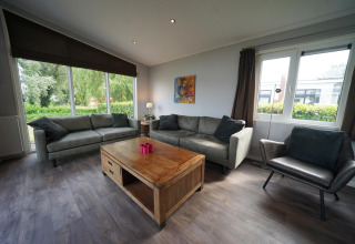 Woonkamer in L-Pavilion 6 bij De Biesbosch, Nederland, met grote ramen, banken en houten tafel.