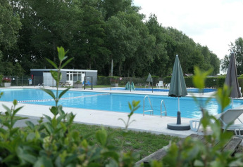 Piscina exterior en L-Pavilion 6 en De Biesbosch, Países Bajos, rodeada de árboles y tumbonas blancas.