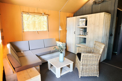 Sala de estar en tienda Safari en Wilsumer Berge, Alemania, con sofá gris, silla de mimbre y muebles de madera.