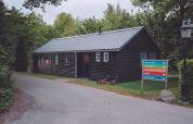 Et sort træhus ved en glampingplads med et farverigt vejskilt og en børnecykel på græsset.