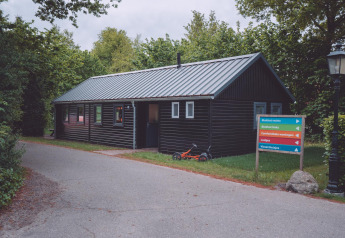 Et sort træhus ved en glampingplads med et farverigt vejskilt og en børnecykel på græsset.
