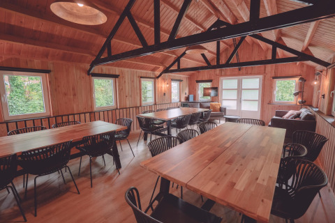Sala comunal rústica con paneles de madera, mesas grandes, sillas negras y sofás en un glamping.