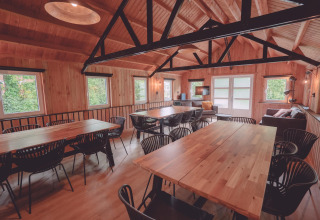 Sala comunal rústica con paneles de madera, mesas grandes, sillas negras y sofás en un glamping.