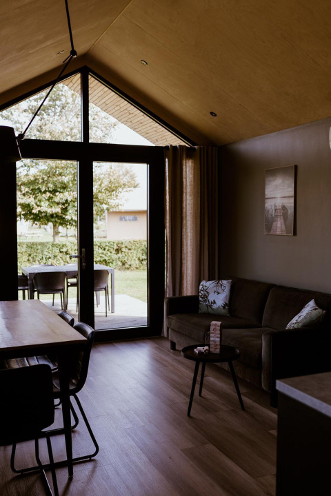 Innenaufnahme einer gemütlichen Lodge mit großen Fenstern, Sofa und Esstisch im Holiday Park Eigen Wijze.
