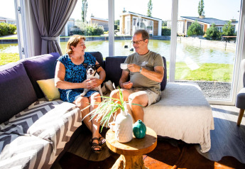 Una pareja mayor y su perro disfrutan café juntos en una sala luminosa con vista al lago en Resort Mooi Bemelen.