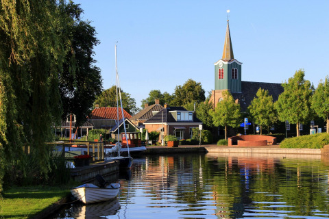 Foto van een gezellige glampingplek aan het water met bootjes, bomen en een kerktoren in het dorp.