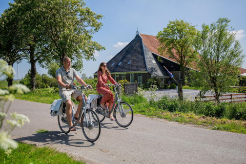 Dos personas pasean en bicicleta junto a un alojamiento de glamping con techo a dos aguas en un día soleado.