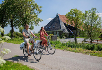 Twee personen fietsen voorbij een charmante glamping-accommodatie met zadeldak op een zonnige dag.