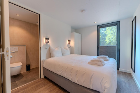 Moderne slaapkamer met tweepersoonsbed, badkamer en groot raam in Forest Cabin, BreeBronne, Nederland.