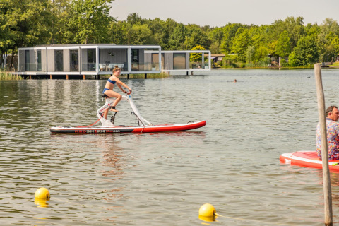 Mujer paseando en una bicicleta acuática frente a cabañas glamping modernas junto al lago y naturaleza.