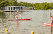 Donna che pedala su una bici d’acqua in un resort glamping sul lago, con alloggi moderni sullo sfondo.