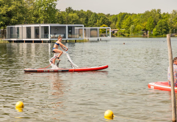 Donna che pedala su una bici d’acqua in un resort glamping sul lago, con alloggi moderni sullo sfondo.