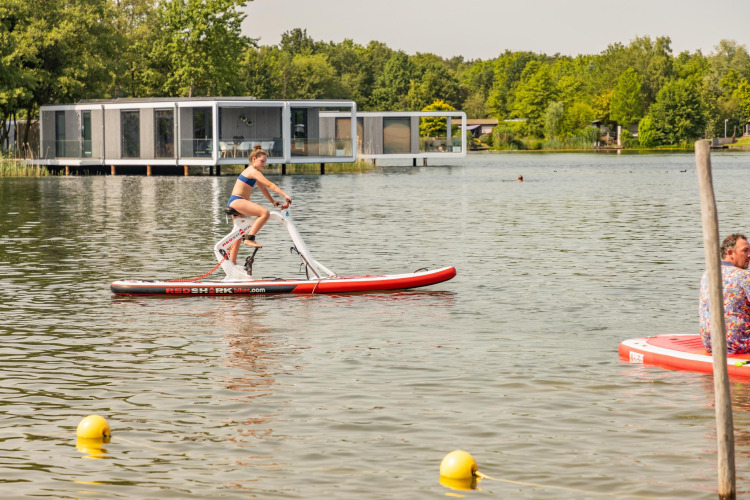 Woman riding a water bike at a lake glamping resort, with modern cabins visible in the background.