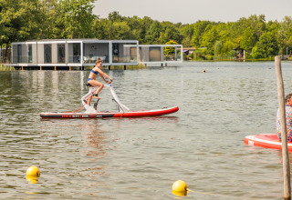 Vrouw fietst op een waterfiets bij een meer, met moderne glamping accommodaties op de achtergrond.