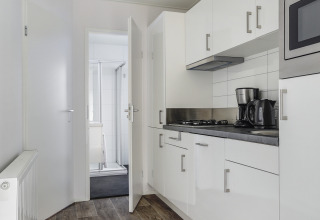 Cucina moderna di tiny house con mobili bianchi, macchina da caffè, bollitore e vista sul bagno.
