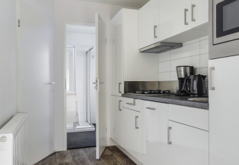 Moderne mikrokøkken i et lille hus med hvide skabe, kaffemaskine, elkedel og udsigt til badeværelse.