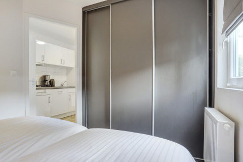 Modernes Schlafzimmer mit grauem Schiebetürenschrank und Blick in die weiße Küche im Tiny House in Holland.