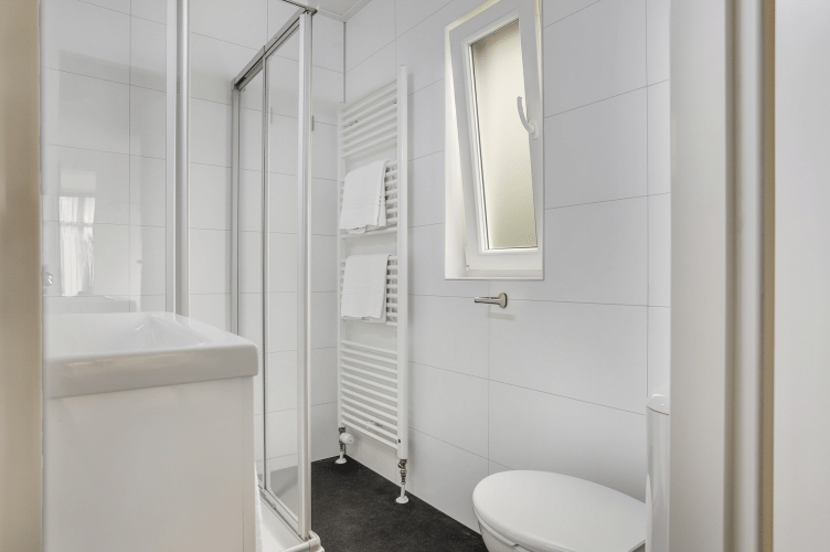 Baño compacto y moderno con ducha, radiador toallero, inodoro y ventana en Tiny House de Bospark Markelo.