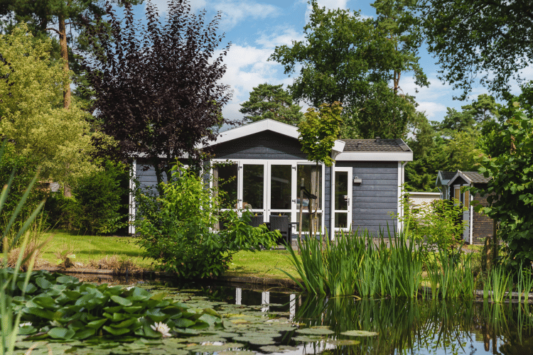 Pequeña casa junto a un estanque rodeada de vegetación en Bospark Markelo, Países Bajos, en un día soleado.