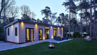 Cubus Lodge in Bospark Markelo, Nederland, moderne houten lodge met terras omgeven door bos in avondlicht.