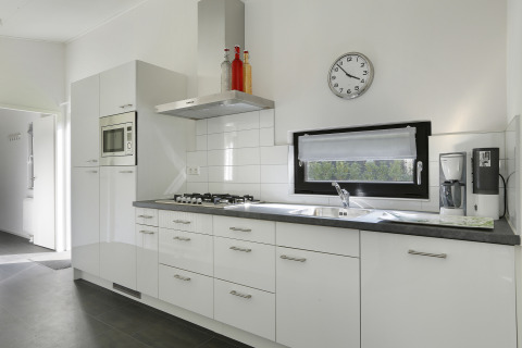 Moderne keuken in Bos Lodge bij ‘t Schuttenbelt met witte kasten, gaskookplaat, microgolfoven en klok.