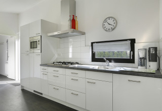 Moderne keuken in Bos Lodge bij ‘t Schuttenbelt met witte kasten, gaskookplaat, microgolfoven en klok.