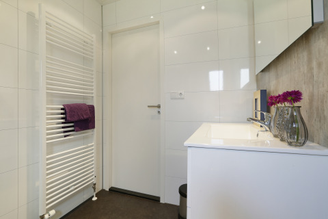 Baño moderno en Bos Lodge, con puerta blanca, toallero calefactor y lavabo con flores moradas en jarrón.
