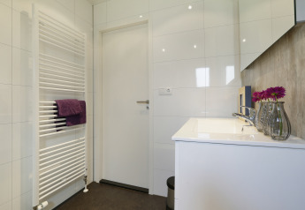 Moderne badkamer in Bos Lodge met witte deur, handdoekradiator en wastafel met paarse bloemen in vaas.