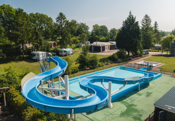 Sistemazione glamping con scivolo d’acqua blu e piscina, circondata da alberi verdi, roulotte e bungalow.