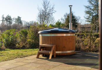 Houten buitenspa met deksel en trapje op betegeld terras bij lodge met uitzicht op de tuin.