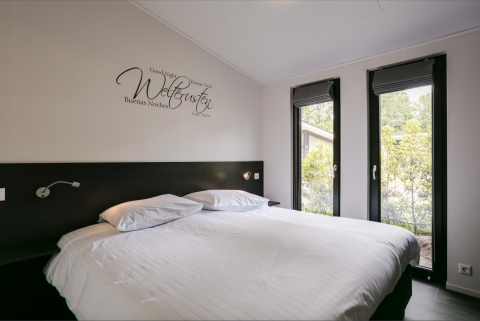 Modern bedroom in Forest Lodge with large bed, wall art and tall windows at Holiday Resort 't Schuttenbelt.