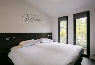 Modern bedroom in Forest Lodge with large bed, wall art and tall windows at Holiday Resort 't Schuttenbelt.