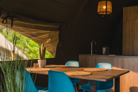 Interior of a safari tent with modern kitchen, wooden table, blue chairs, and a window to green trees.