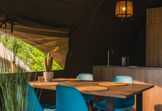 Interior of a safari tent with modern kitchen, wooden table, blue chairs, and a window to green trees.