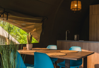 Interieur van een safaritent met moderne keuken, houten tafel, blauwe stoelen en uitzicht op bomen.