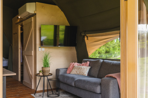 Interior of a safari tent lodge with a grey sofa, TV, and potted plant at Holiday Resort 't Schuttenbelt, Netherlands.