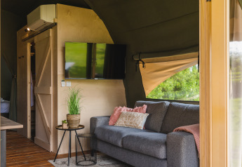 Intérieur d'une tente lodge safari XL avec canapé gris et télé à Holiday Resort 't Schuttenbelt, Pays-Bas.