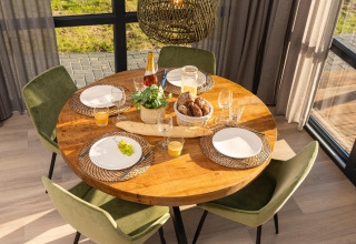 Runder Holztisch im Sonnenschein, gedeckt für vier Personen mit Brot, Saft, Wein und Zierpflanze.