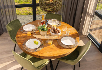 Ronde houten eettafel gedekt voor vier, met brood, sinaasappelsap, wijn en een plant in zonlicht.